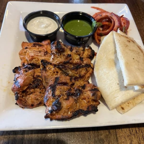 Grilled chicken with two dipping sauces, pickled onions, and pita bread on a white plate.